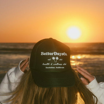 Person holding a black “BetterDaysCo.” hat at the beach during sunset, with the sun over the ocean.