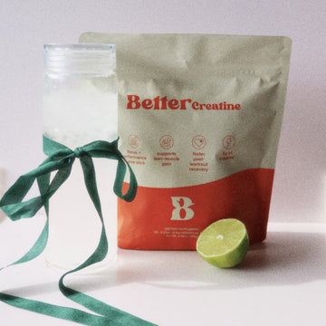 Pouch labeled “Better Creatine” beside a clear bottle with green ribbon and a halved lime on a white surface.