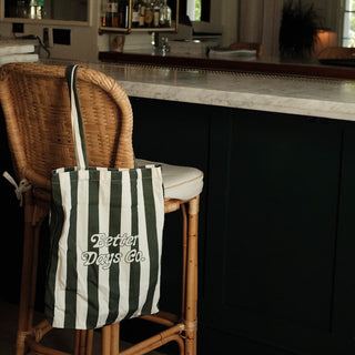 Striped tote bag reading “Better Days Co.” hanging on a wicker barstool beside a marble countertop.