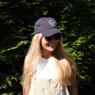 Smiling woman with long blonde hair wearing a navy cap and sunglasses, standing in front of green foliage.