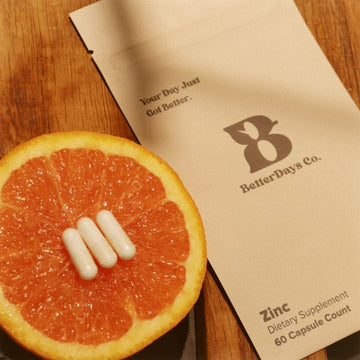 Half grapefruit with three white capsules on top beside a BetterDays Co. zinc supplement packet on a wooden surface.