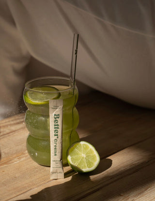 Glass of green drink with lime slices and a 'Better Greens' powder stick on a wooden surface.