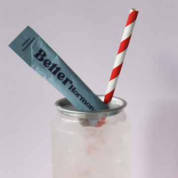 Can with a red-and-white striped straw and a blue “Better Hormone” supplement packet against a pale background