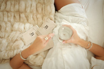 Person lying on bed, holding a glass, with two product boxes on a beige furry blanket.