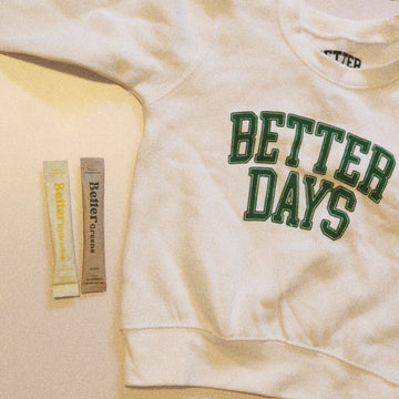 White sweatshirt with green “BETTER DAYS” text, next to two BetterGreens packets on a beige surface.