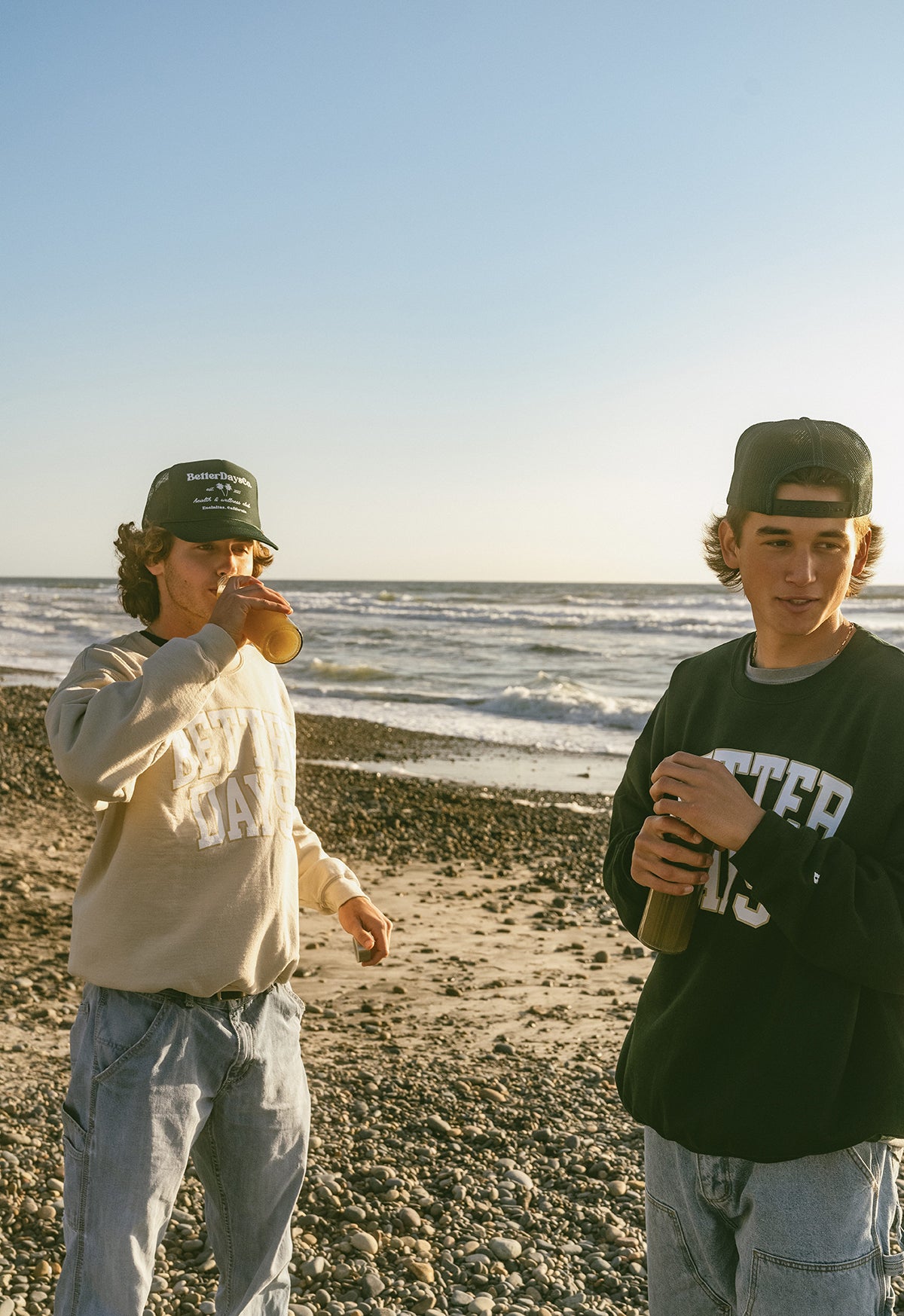 Two people in sweatshirts and caps holding drinks on a rocky beach with ocean waves behind them.