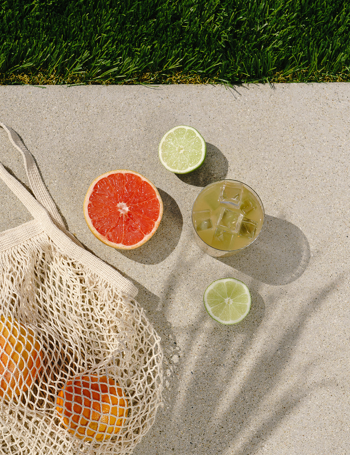 Mesh produce bag with oranges, half grapefruit, lime halves, and a glass of iced citrus drink on concrete by grass.