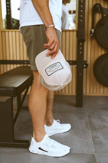 Person in shorts holding a white baseball cap in a gym, standing beside a bench and weight rack.