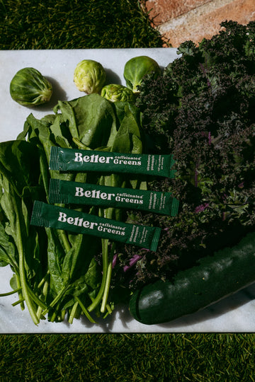 Three Better Greens packets on leafy greens with kale, Brussels sprouts, and a zucchini on a cutting board.