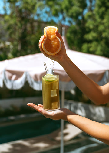 Hand squeezing a grapefruit half over an iced drink in a tall glass with a straw and lime slice outdoors.