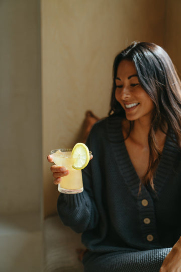 Smiling woman holding a glass of lemon drink with a lemon slice garnish indoors.
