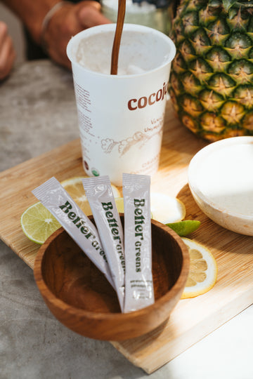 Wooden bowl with BetterGreens packets, sliced citrus on a cutting board, coconut yogurt cup being stirred, and a pineapple.