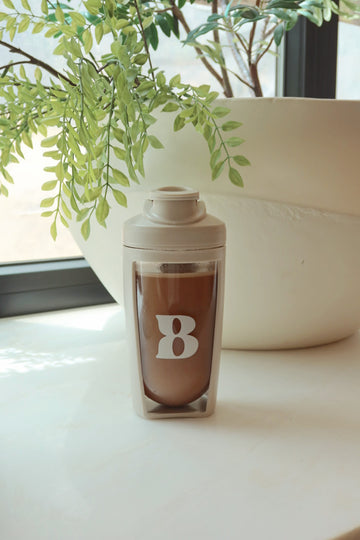 Gray shaker bottle with brown drink and white “8” logo on a table beside a leafy potted plant.