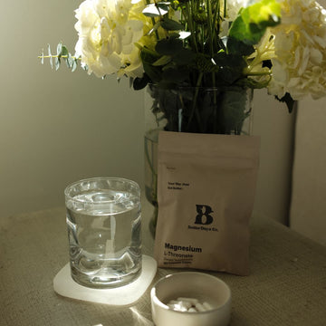 Glass of water on a coaster beside a magnesium supplement pouch, with a vase of white flowers in background.