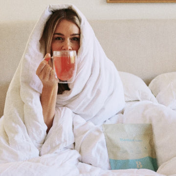 Person in bed wrapped in a white blanket, holding a clear mug with a pink drink.