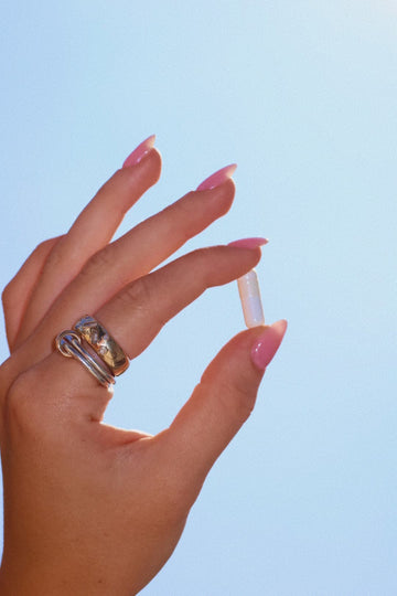 Hand with pink manicured nails and a silver ring holding a clear capsule against a blue sky.
