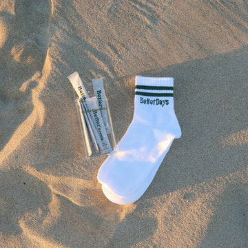 White sock with green stripes and “BetterDays” text beside a clear pouch of packets on sand.