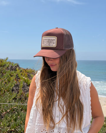 A woman wearing a brown and burgundy trucker hat with a beige 'BITTERDAYS' patch on the front, photographed outdoors against a coastal background with ocean and sky.