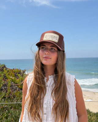 A woman with long light brown hair stands outdoors on a beach dune wearing a brown baseball cap with a beige patch that reads 'BETTER DAYS' and a white sleeveless top. The background shows the ocean, sky, and sandy beach.