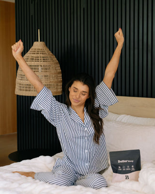 A woman wearing a blue and white striped pajama set sits on a bed with her arms raised in the morning, next to a black pouch labeled 'Better Sleep'.