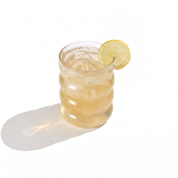 A ribbed glass tumbler of pale gold juice over ice, garnished with a lemon wheel on the rim, on a white background