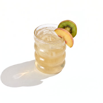 A ribbed glass tumbler of pale gold juice over ice, garnished with a peach slice and a kiwi half on the rim, on a white background