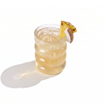 A ribbed glass tumbler of pale gold juice over ice, garnished with a pineapple wedge and a coconut chunk on the rim, on a white background