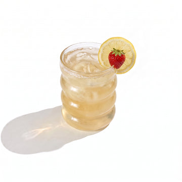 A ribbed glass tumbler of pale gold juice over ice, garnished with a strawberry and a lemon wheel on the rim, on a white background