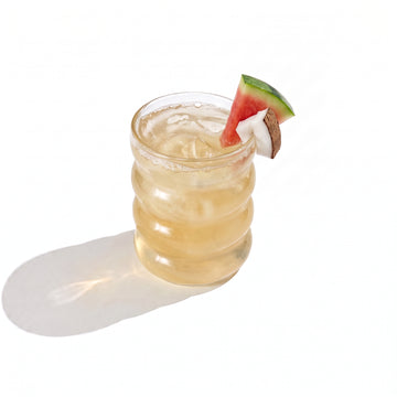 A ribbed glass tumbler of pale gold juice over ice, garnished with a watermelon wedge and a coconut chunk on the rim, on a white background