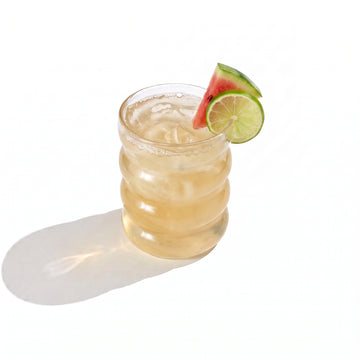 A ribbed glass tumbler of pale gold juice over ice, garnished with a watermelon wedge and a lime wheel on the rim, on a white background