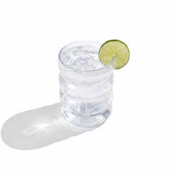 A ribbed glass tumbler of clear sparkling water over ice, garnished with a lime wheel on the rim, on a white background