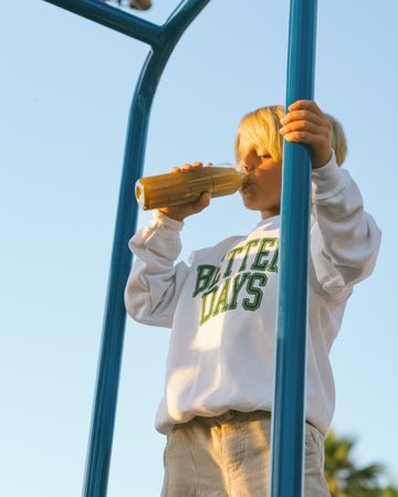 Child in a white sweatshirt drinking from a bottle while holding a blue playground pole.
