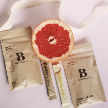 Half grapefruit on white fabric beside BetterDays Co. supplement packets and stick packs labeled Zinc and Vitamin D3+K2.