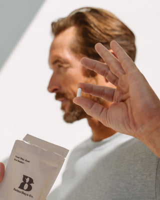 A man in a gray t-shirt holds a white blister pack and a single white tablet between his fingers, shown in a close-up profile against a bright, minimalist background.